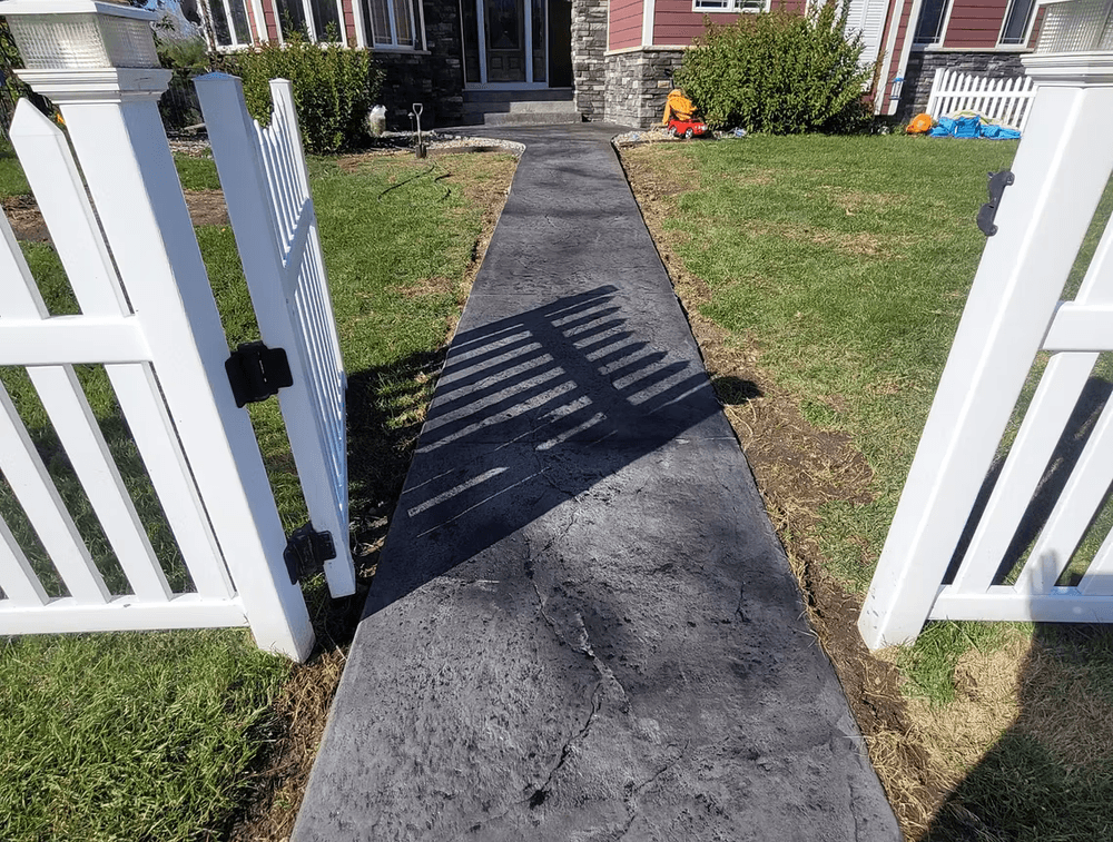 Concrete walkway leading to a house, bordered by a white picket fence and a manicured lawn.