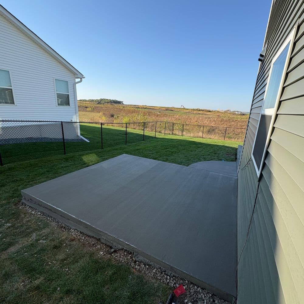 Freshly poured concrete patio beside a house with a fenced yard and open landscape.