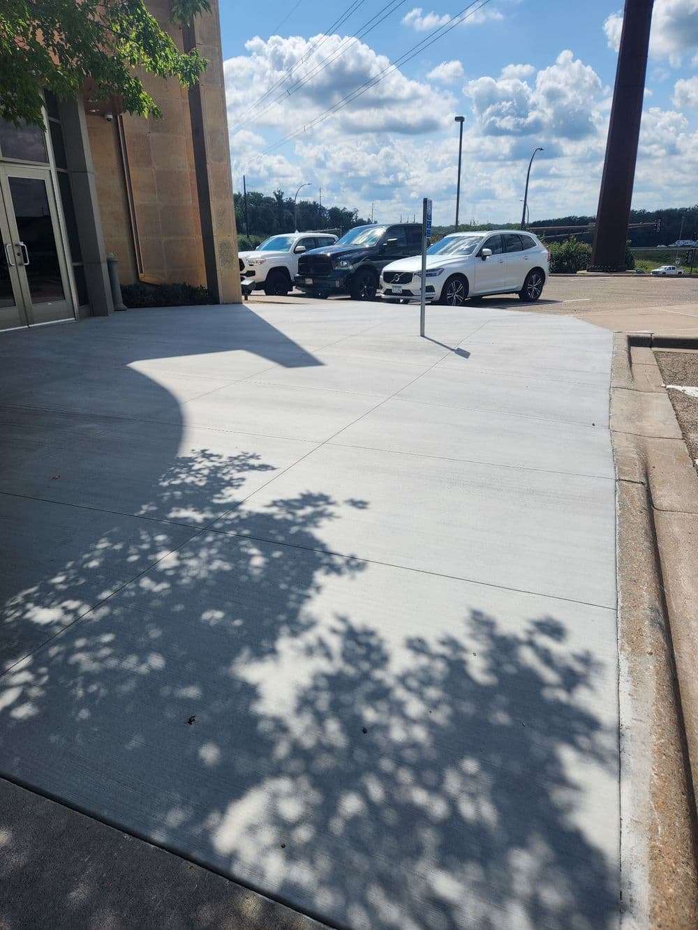 Concrete sidewalk with shadows and parked cars under a sunny sky.