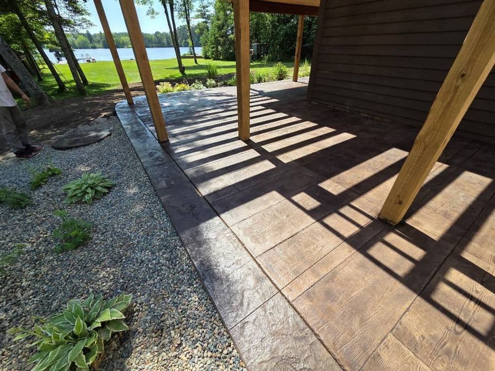 Patio with stamped concrete, wooden beams, and landscaping near a lake under shaded sunlight.