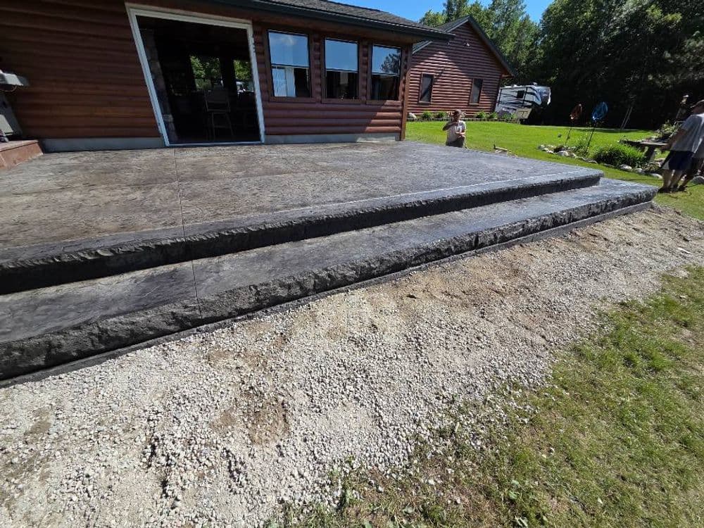 Concrete patio with steps, surrounded by grass and a wooden house in the background.