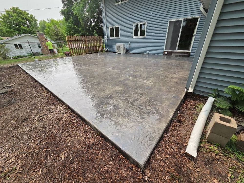 Newly poured concrete patio beside a blue house with an air conditioner and landscaped area.