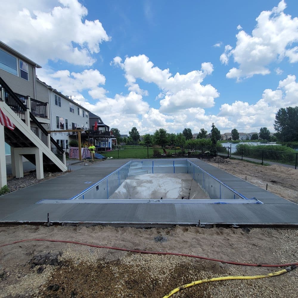 In-ground pool construction site with concrete base and surrounding landscaping, under a blue sky.
